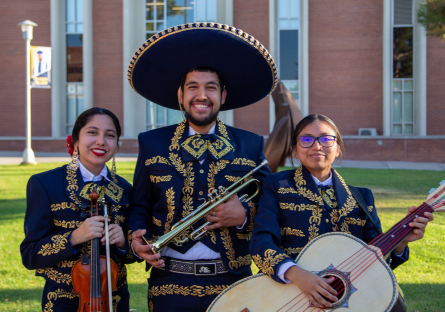 Three Phoenix College Mariachi Ensemble members stand on campus in traditional trajes with their instruments