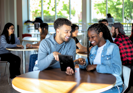 two students look at a tablet and smile in a crowded cafeteria