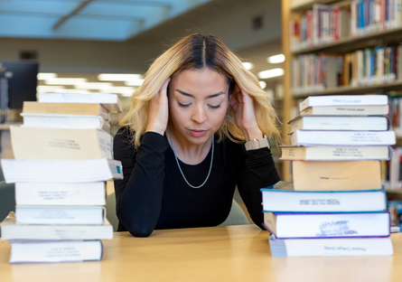 female student holds head in sadness next to piles of books