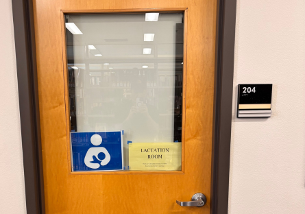 Image of Library Lactation Room Door with blue sign and white icon of an adult holding an infant with additional yellow sign that reads "Lactation Room"