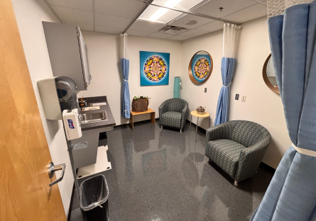 Interior of Nursing Center Lactation Room with a small sink and upper cupboards on the left wall and two gray upholstered chairs on the opposite wall with a small round table between them and colorful artwork on the far wall. 