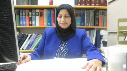 PC engineering professor Dr. Amal Saeed Yagub sits at her desk looking at papers on her desk with a bookcase of books behind her