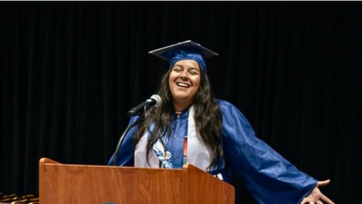 Phoenix College graduate Ivonne Dominguez in her cap and gown at a podium speaking with her arms outstretched