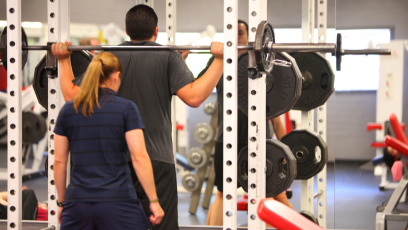 A man performs weighted squats on a Smith machine supervised by a female fitness coach in Phoenix College's Fitness Center 