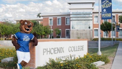 Phoenix College Mascot standing next to Phoenix College sign on campus with Fannin Library in background