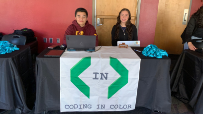 Kenneth Ho and Shelby Alonso sit at a table with a Coding in Color banner ready to welcome high school student to the Day of Compute at Phoenix College
