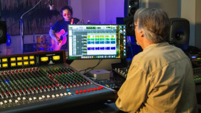 Musician Troy Lowney stands behind a microphone in a soundbooth with a guitar in his hands while PC student Joe Drzewiecki sits at an audio mixing board in the foreground