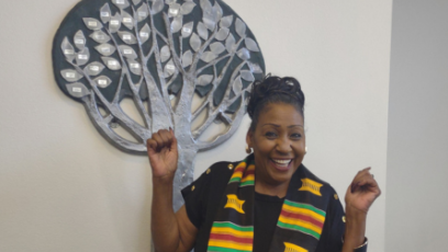 PC Psychology Professor Dr. Debbie Webster stand in front of the Black Student Union Living Legacy Award Tree in Fannin Library wearing a black top and black stole with green yellow and red stripes