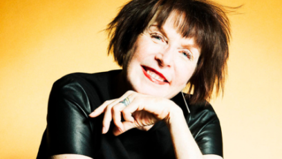Artist Marilyn Minter wears a black top with a bright yellow background, with her left hand resting under her chin, smiling. 