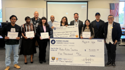 Students who competed in the Bear's Den competition stand in a classroom with a projection screen holding certificates and large checks in front of them with competition organizers and judges standing with them