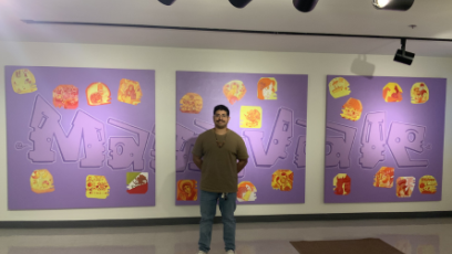 Edgar Fernandez stand in front of the three-panel mural he created with art students from Maryvale High School. The mural has a purple background with corn kernels reflecting the students identities in shades of yellow, red, and orange. 