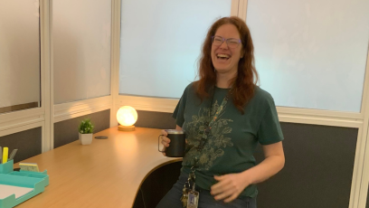 Phooenix College Learning Commons Manager, Dr. Sam Crandall, smiles and stands at an adjustable desk with a small warm light circle lamp to show off some of the features of the study rooms she built for neurodivergent students.  
