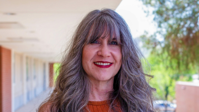PC Education Director Kat Valdivia stand outsides the 2nd floor of the H building where she works, smiling with her salt and pepper bangs in her eyes and her long hair past her shoulders 