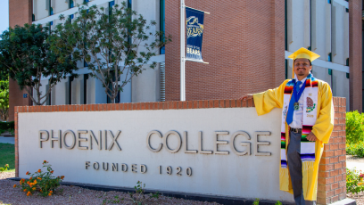 Luis Duque stand in his graduation cap and gown next to the sign on Thomas Road: "Phoenix College Established 1920"