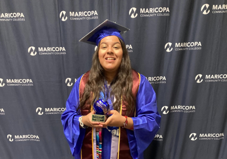 Phoenix College graduate Ivonne Dominguez in a blue cap and gown stands in front of a Maricopa County Community College District step and repeat backdrop holding an award