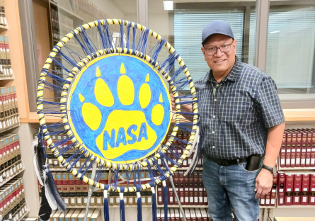 Phoenix College Native American Student Association (NASA) President Melvin Pastores stands next to the NASA logo, a bear claw inside a dream catcher hoop.