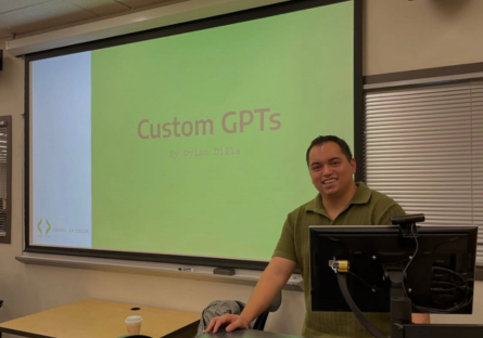 Coding in Color instructor Dylan Dilla stands at an instructor computer station with his intro slide "Custom GPTs" with a green background projected on the screen.