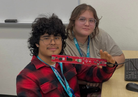 Carl Hayden high school students Diego Diaz and Rose Franco, sit in a Phoenix College computer lab during Day of Compute with Diego holding up a long, narrow circuit board in his left hand.