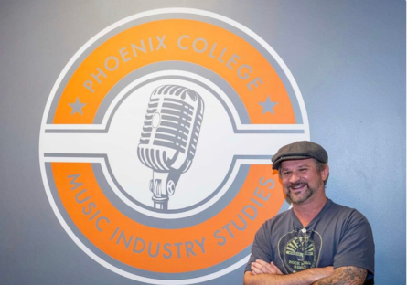 Jamison Weddle, PC Music Industry Program Director, stands next to the gray and orange logo for the Music Industry Studies logo with a vintage microphone in the center
