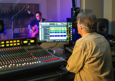Musician Troy Lowney stands behind a microphone in a soundbooth with a guitar in his hands while PC student Joe Drzewiecki sits at an audio mixing board in the foreground