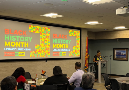 Dr. Webster stands at a podium in the Hacienda room with a slide with words "Black History Month Legacy Luncheon February 25th" projected on the screen with the audience sitting at round tables