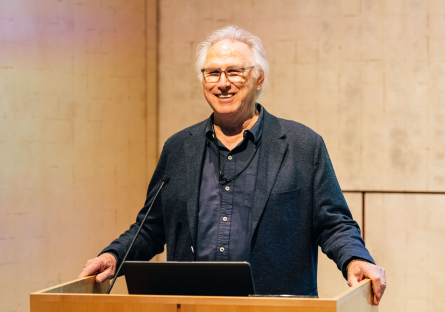 Aritst Eric Fischl stands at the podium in Phoenix Art Museum's Whiteman hall with gold walls, wearing a dark blue shirt and jacket, his hand resting on the side of the podium 