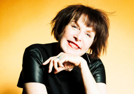 Artist Marilyn Minter wears a black top with a bright yellow background, with her left hand resting under her chin, smiling. 