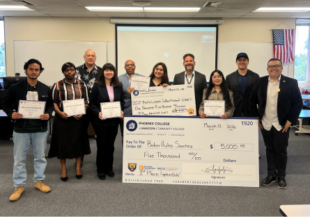 Students who competed in the Bear's Den competition stand in a classroom with a projection screen holding certificates and large checks in front of them with competition organizers and judges standing with them