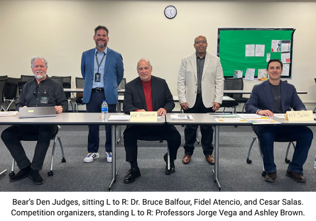 Bear's Den competition judges Dr. Bruce Balfour, Fidel Atencio, and Cesar Sals sit at a long table with competition organizers Professors Jorge Vega and L. Ashley Brown standing in suits behind the judges in a classroom with a clock and bulletin board visible in the background.  