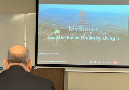 A projection screen shows a slide with a landscape image of rural Italy with the words "MyBorgo: Save the Italian Dream by Living it" with the backside of a judges head and shoulder visible in the foreground. 