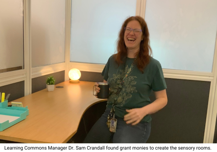 Phooenix College Learning Commons Manager, Dr. Sam Crandall, smiles and stands at an adjustable desk with a small warm light circle lamp to show off some of the features of the study rooms she built for neurodivergent students.  