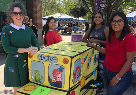 Phoenix College Education Director Kat Valdivia is dressed as Ms. Frizzle in a green dress with white collar and pins of planets pinned to the dress. She stands next to a cardboard vehicle with the words "Blast Off" on the front with future teachers in PC's Education program.