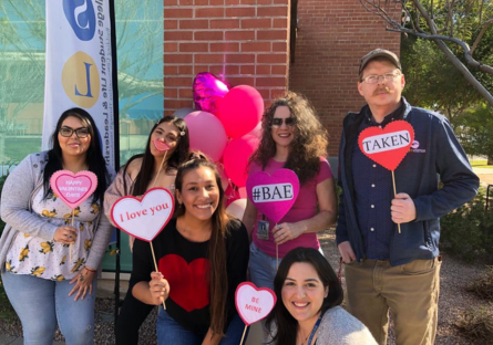 Phoenix College Education Director Kat Valdivia stands with students in the Education program holding heart signs with text in the middle of the hearts reading "#BAE", "I love you", "Be Mine", "Taken" and surrounded with heart ballons during the Valentine Day Marketplace supporting the Future Teacher Club at Phoenix College 