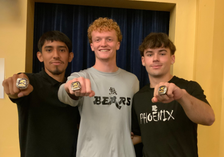 Cruzerio Cruz, Jack Cameron, Landon Vree stand side by side in the Bulpitt auditorium with one fist outstretched to show off their championship rings. 