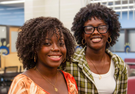 Sister Shadé and Sy'nai Ajayi stand side by side, smiling, in Phoenix College's Learning Commons where they both work while attending school for Medical Assisting and Elementary Education respectively