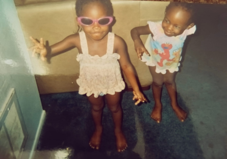 Sy'nai and Shadé Ajayi as small children with Sy'nai wearing pink sunglasses, holding up her right hand trying to make a peace gesture with Shadé looking over to Sy'nai and trying to make the same gesture