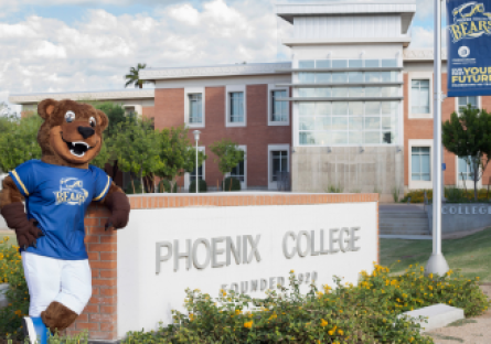 Phoenix College Mascot standing next to Phoenix College sign on campus with Fannin Library in background