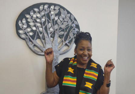 PC Psychology Professor Dr. Debbie Webster stand in front of the Black Student Union Living Legacy Award Tree in Fannin Library wearing a black top and black stole with green yellow and red stripes