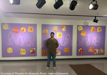 Edgar Fernandez stand in front of the three-panel mural he created with art students from Maryvale High School. The mural has a purple background with corn kernels reflecting the students identities in shades of yellow, red, and orange. 