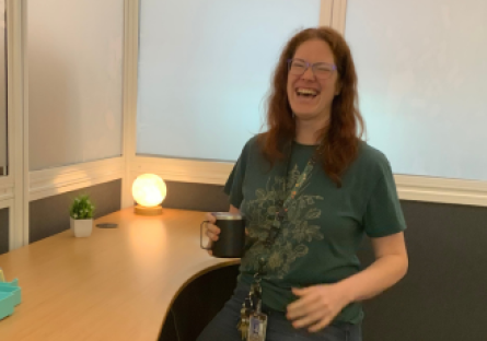 Phooenix College Learning Commons Manager, Dr. Sam Crandall, smiles and stands at an adjustable desk with a small warm light circle lamp to show off some of the features of the study rooms she built for neurodivergent students.  