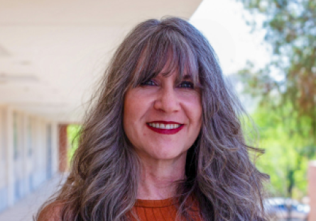 PC Education Director Kat Valdivia stand outsides the 2nd floor of the H building where she works, smiling with her salt and pepper bangs in her eyes and her long hair past her shoulders 