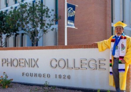 Luis Duque stand in his graduation cap and gown next to the sign on Thomas Road: "Phoenix College Established 1920"