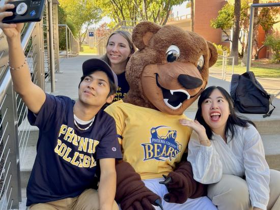 Three students sitting on steps outside taking a selfie with college mascot