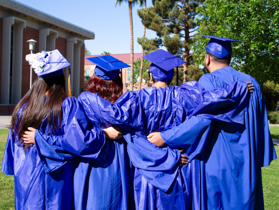 Four students in blue regalia facing away from the camera with arms around each other in Pastor Plaza on PC's campus